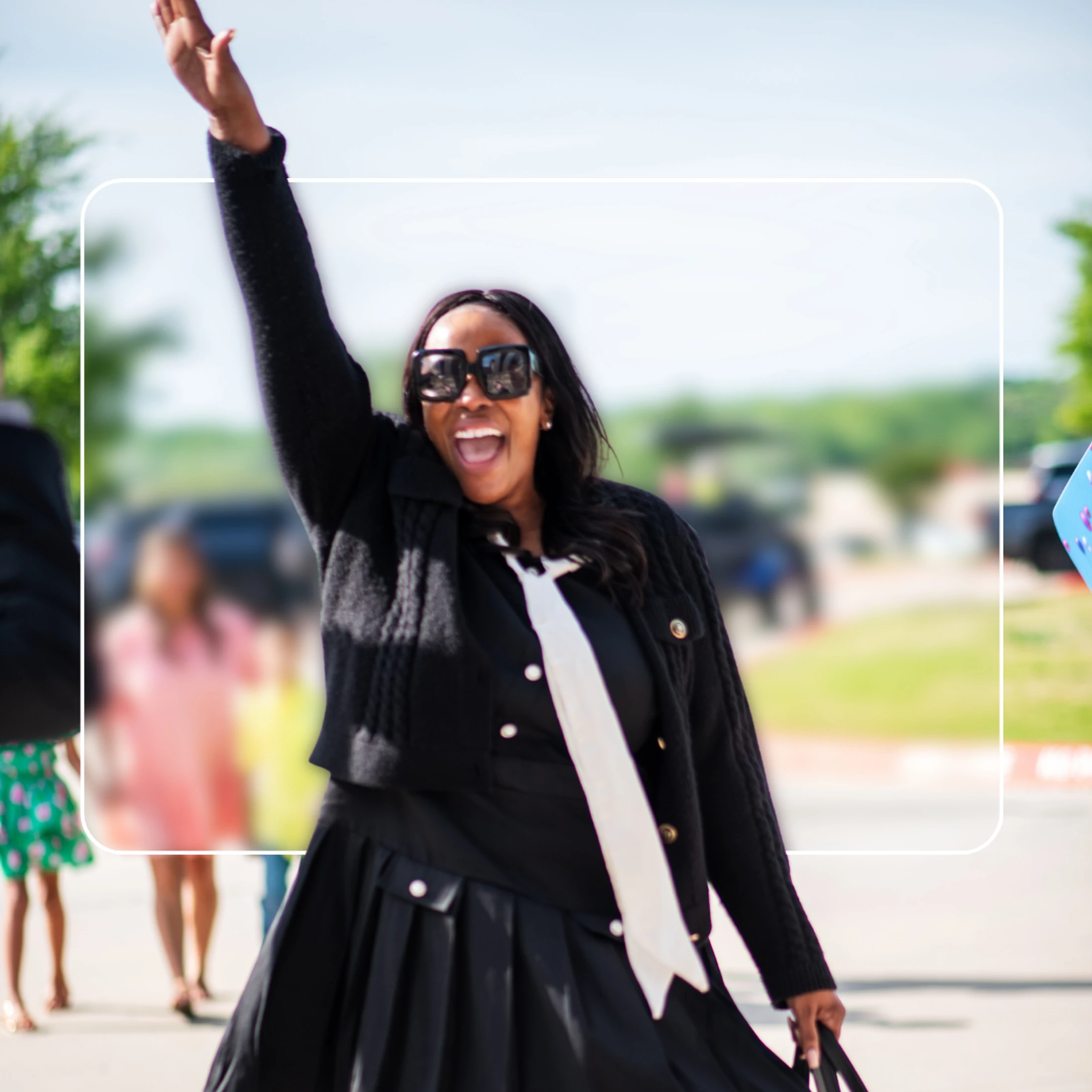 A happy Black woman in sunglasses and a stylish black outfit waves her hand in the air while smiling with her mouth open outdoors on a sunny day.
