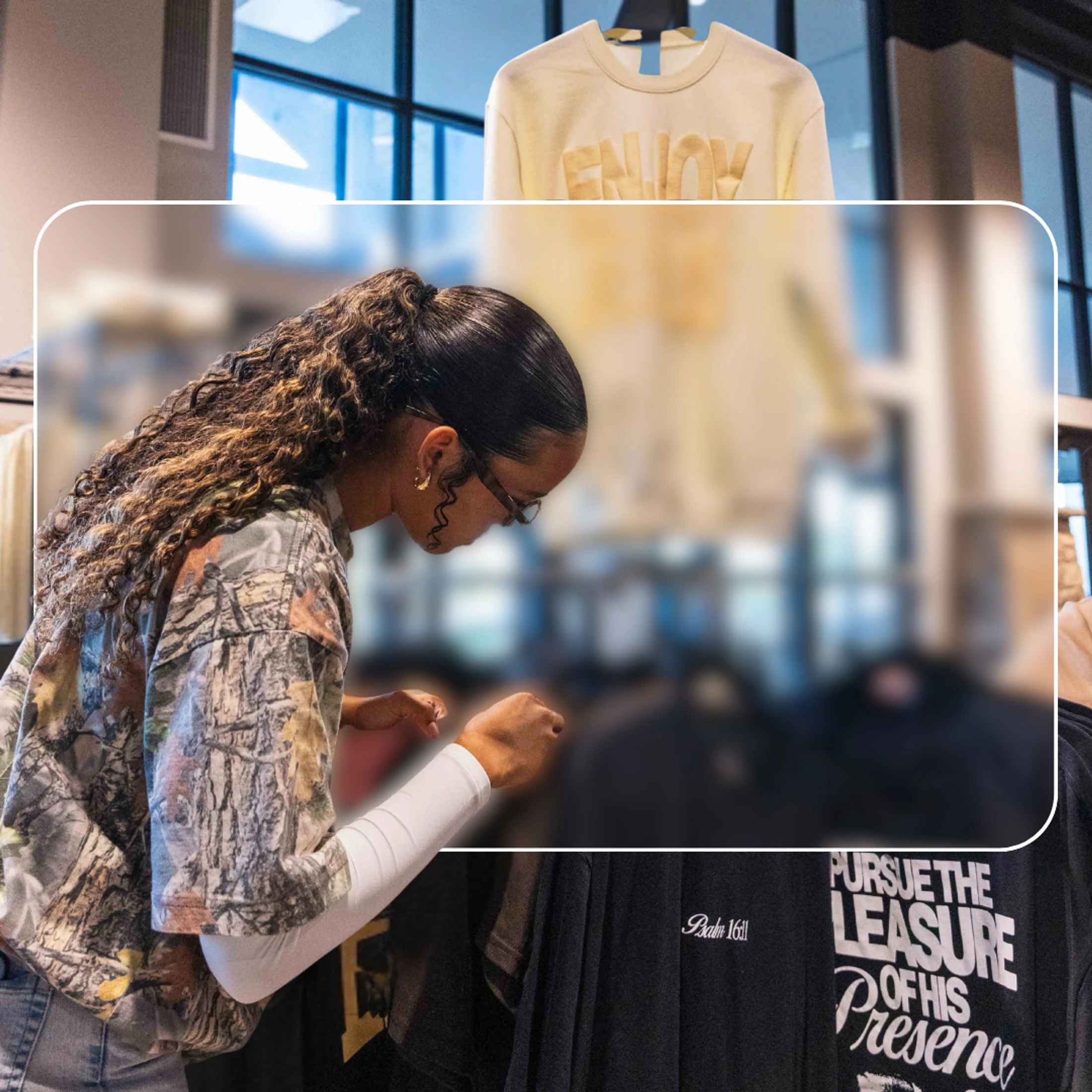 A woman with long curly hair and glasses, wearing a camouflage shirt, looks down at a rack of clothing in a retail shop.