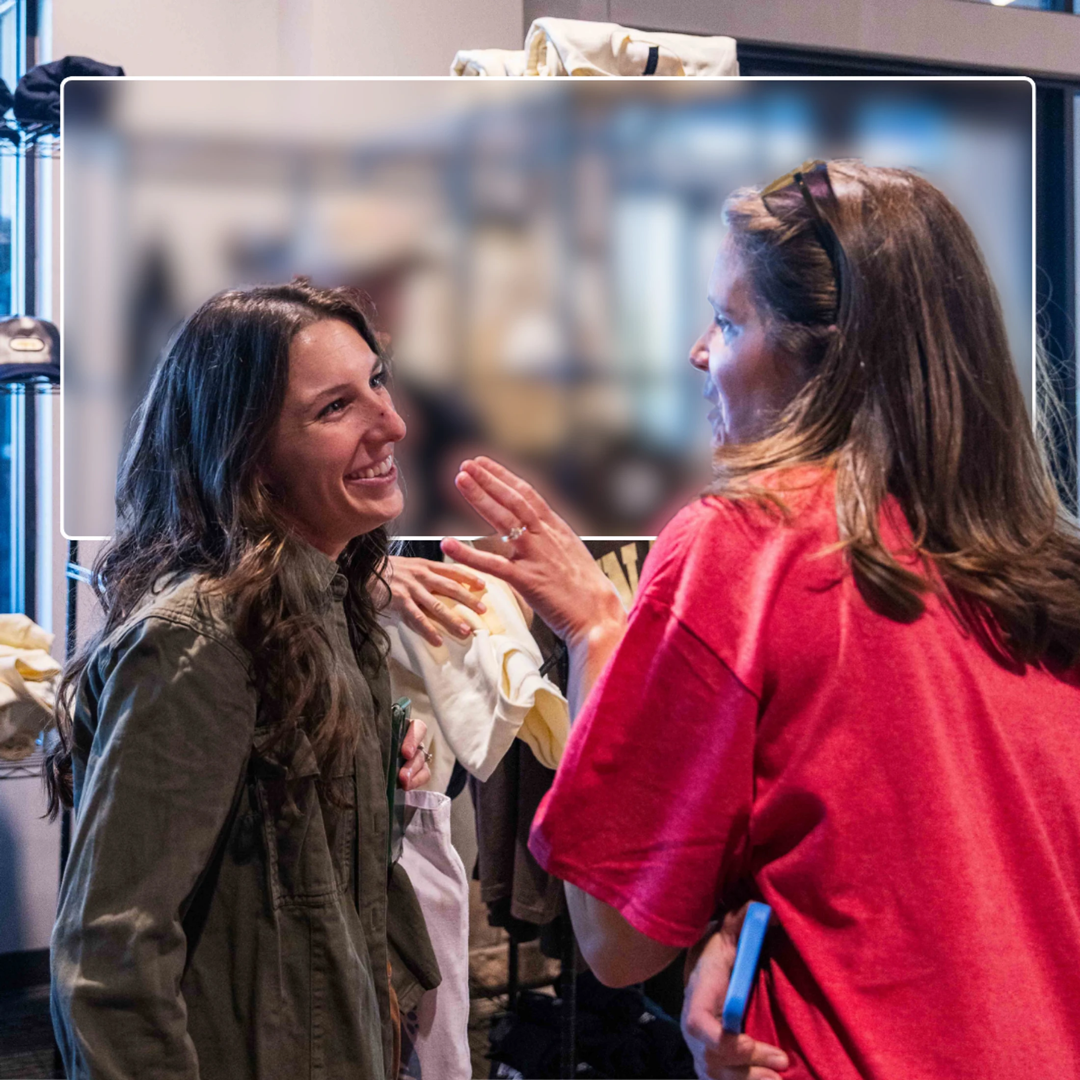 Two women stand talking in a room with clothes racks. The woman on the left smiles as the woman on the right, wearing a red shirt, gestures while speaking.