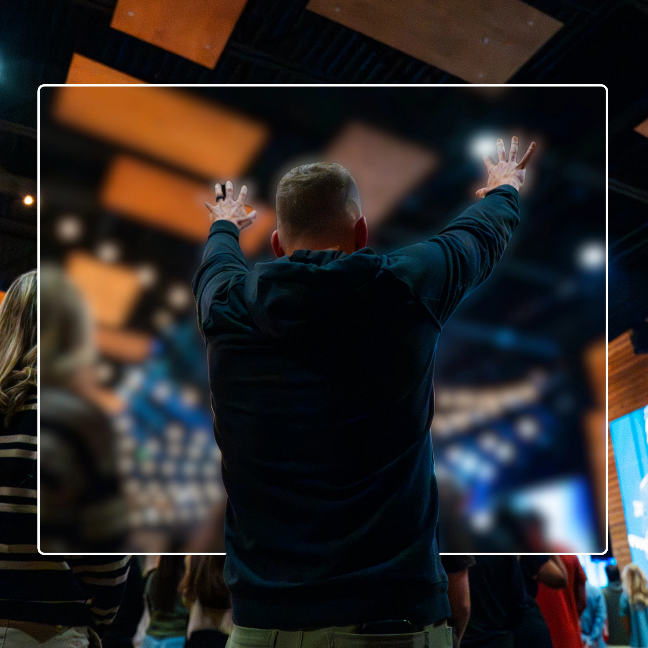 A man viewed from behind in a dark hoodie stands in a crowd with his arms raised high in a gesture of praise during a worship service.
