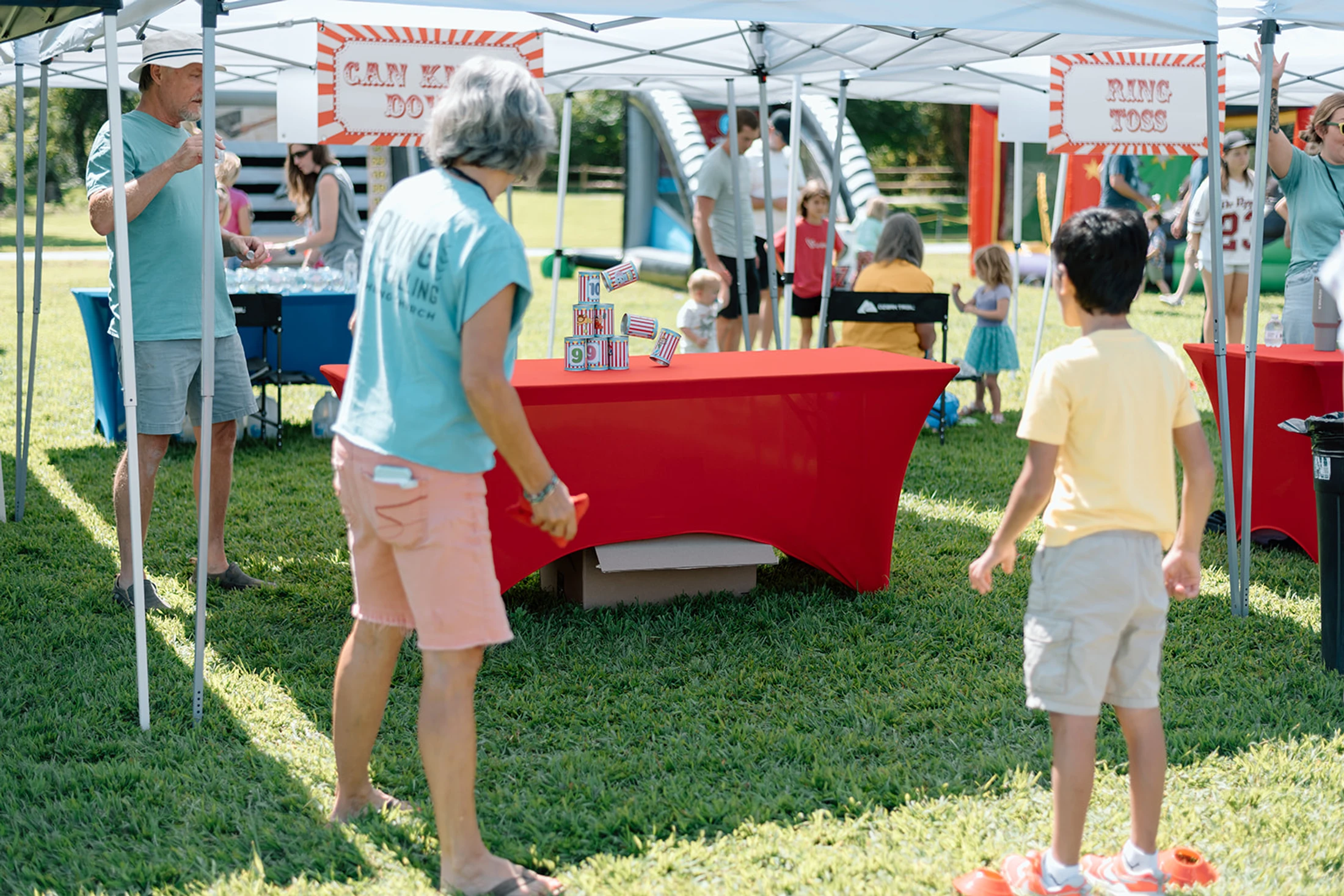 A woman and a young boy stand by a can knockdown game under a white tent at a sunny outdoor festival with other families in the background.