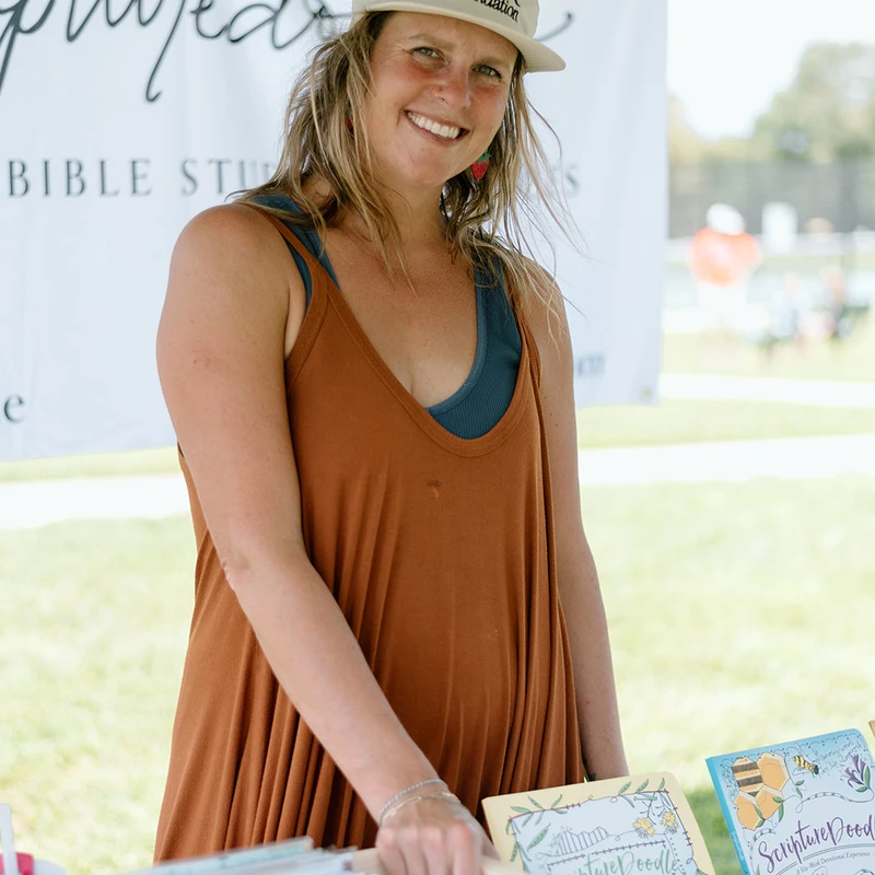 A smiling woman wearing a white hat stands behind a table at an outdoor market, with religious-themed books and cards displayed for sale in a wooden crate.