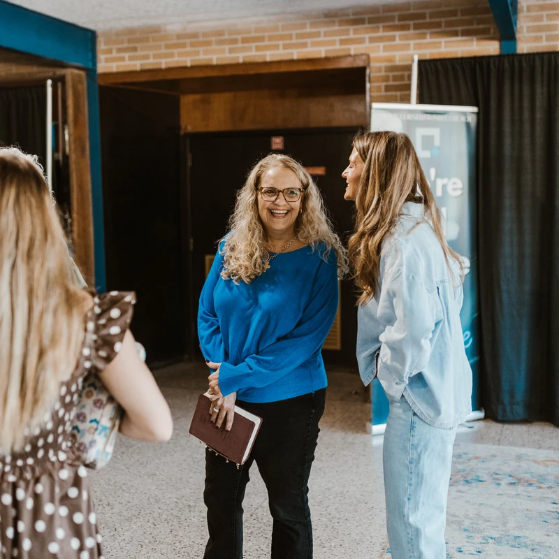 Three women stand in a building's lobby, talking and laughing. A woman in a blue sweater and glasses smiles brightly while holding a book.