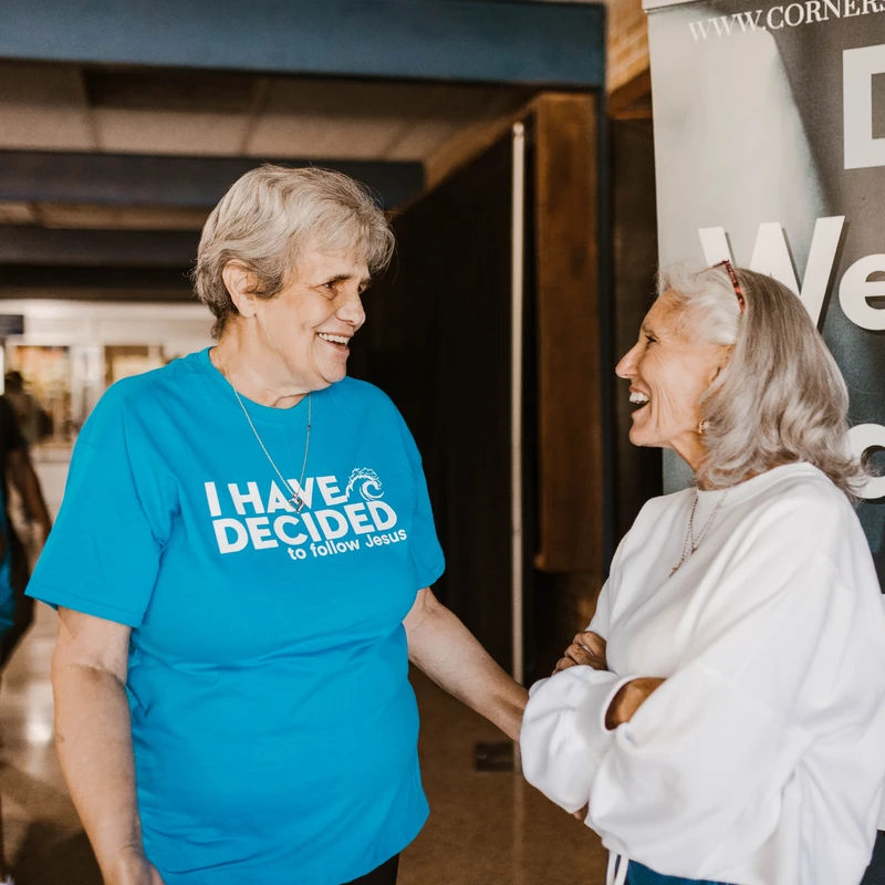 Two older women smiling and talking in a church lobby. One wears a blue t-shirt that reads 'I have decided to follow Jesus' in white letters.