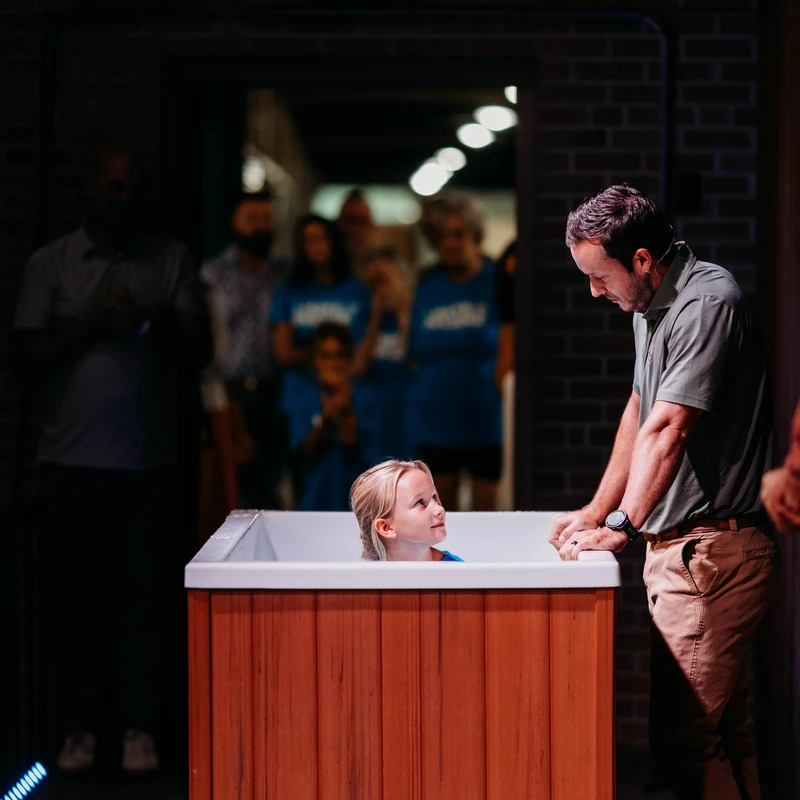 A young blonde girl sits in a portable baptismal font, looking up at a pastor who leans over to speak with her before the religious ceremony begins.