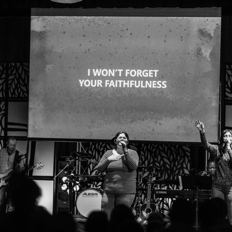 A black and white photo of a worship band on stage. Singers lead worship while a screen behind them says, 'I WON'T FORGET YOUR FAITHFULNESS.'