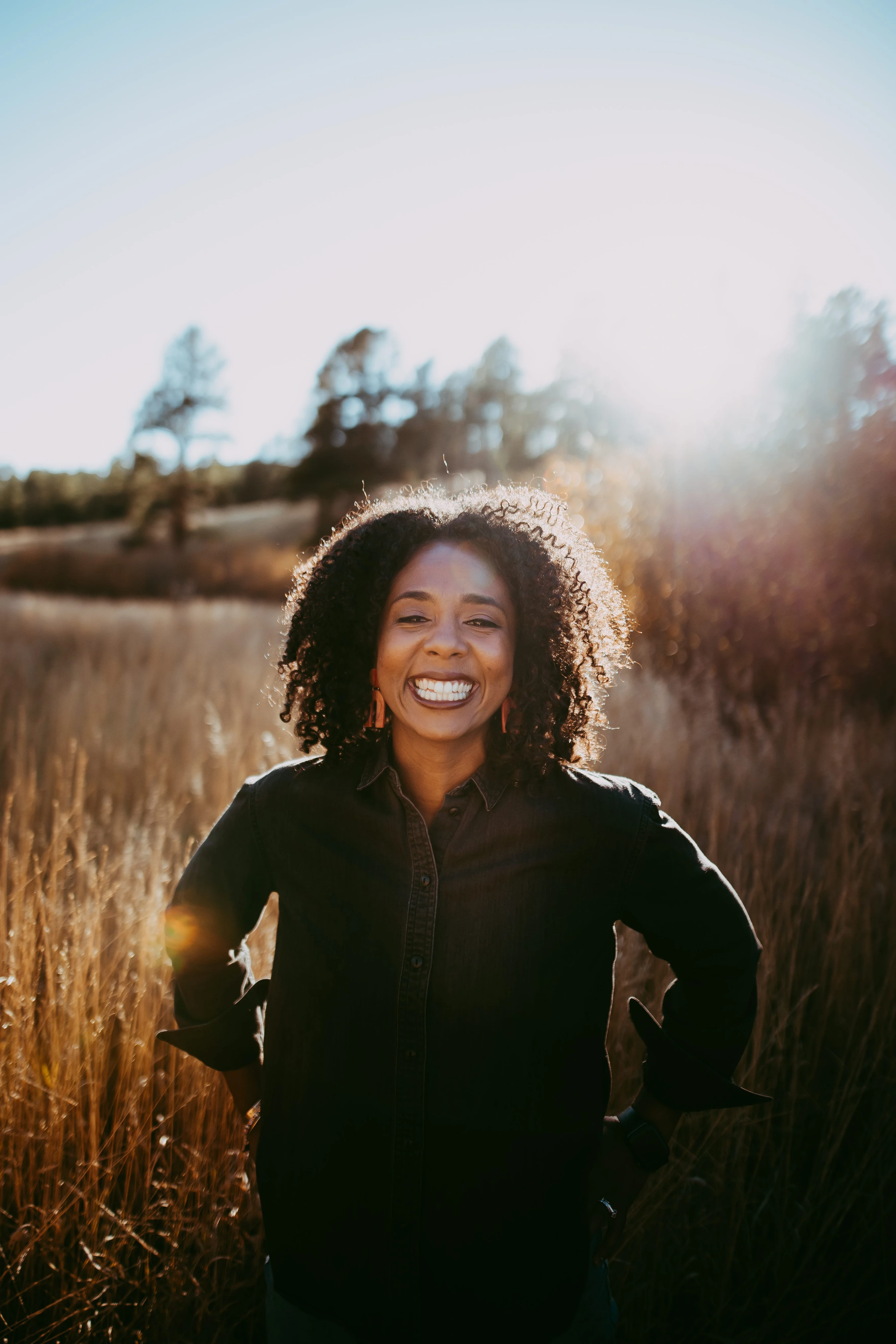 A smiling woman with curly hair, backlit by the setting sun, stands in a field of tall golden grass with her hands on her hips.