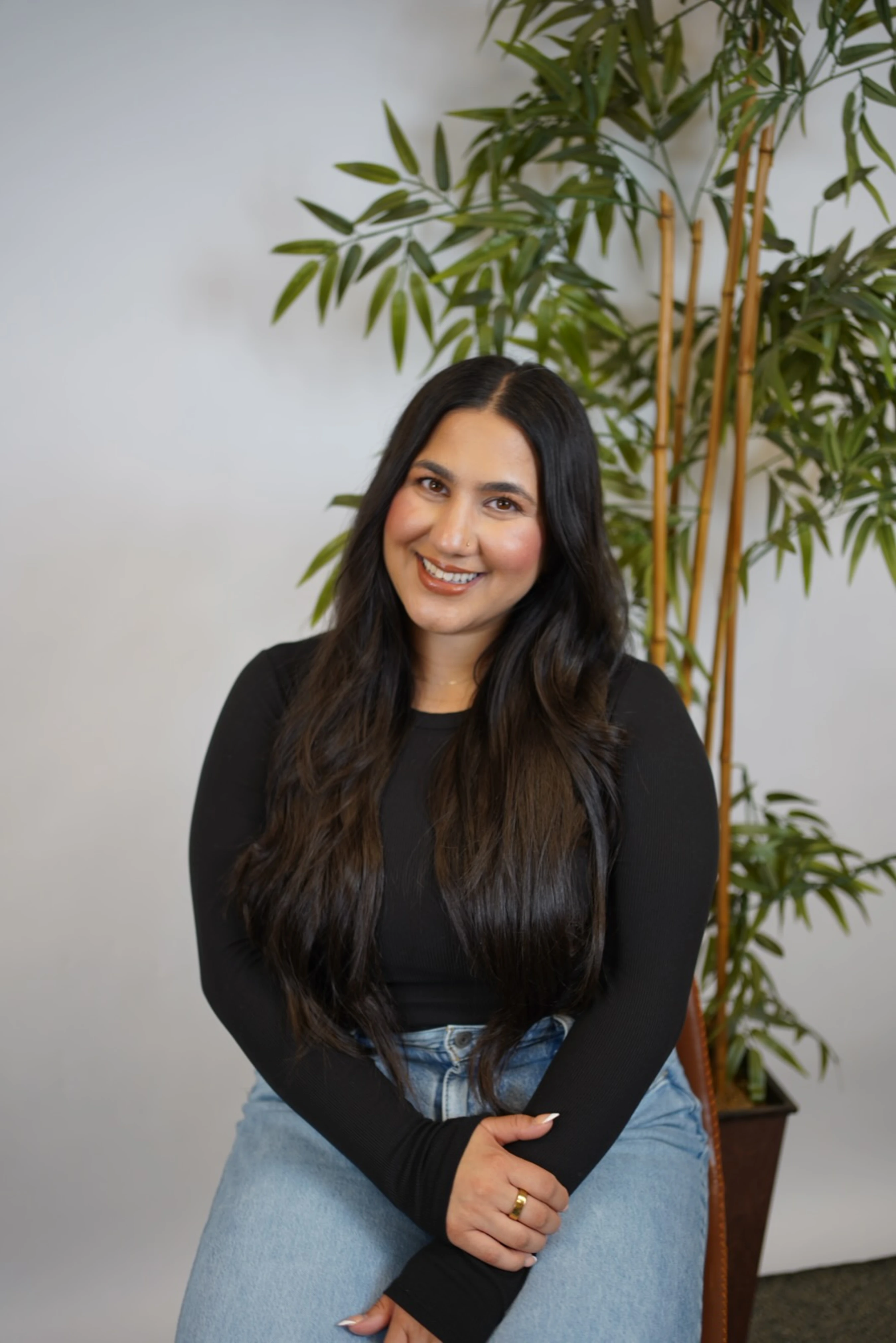 A smiling woman with long dark hair sits for a portrait. She wears a black long-sleeved shirt and jeans, posing in front of a white wall and a tall plant.