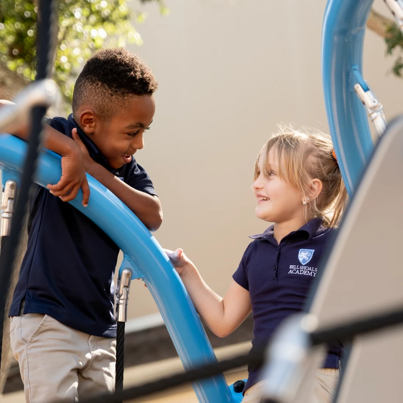 A young Black boy and a young white girl, both in school uniforms, smile at each other while climbing on a blue outdoor playground structure.