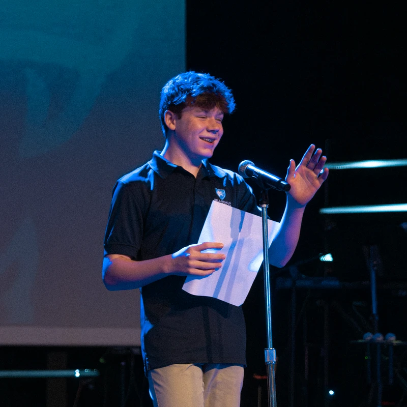 A young male student in a polo shirt smiles while speaking into a microphone on a dimly lit stage, holding a script and gesturing with one hand.