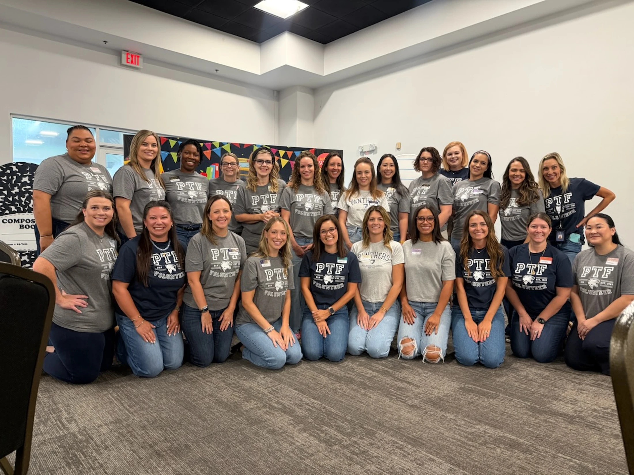 A large group of smiling women, all wearing matching 'PTF Volunteer' t-shirts in grey or navy blue, pose for a group portrait in a room.