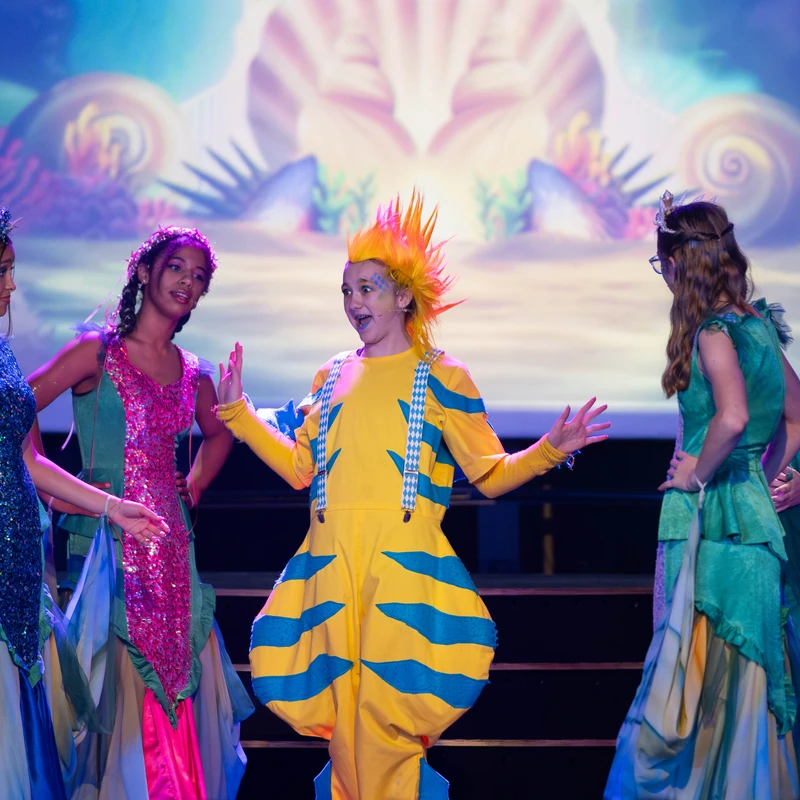 A young actor in a yellow and blue fish costume sings on stage, surrounded by other performers in mermaid-like dresses during a play.
