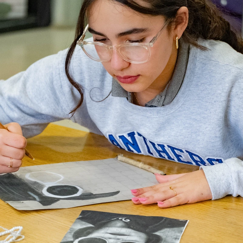 A female student with glasses wearing a grey sweatshirt leans over a desk, carefully drawing a portrait from a black and white photo using the grid method.
