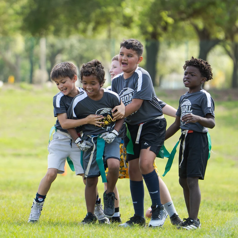 A group of diverse young boys in matching jerseys smile and laugh while playing flag football on a sunny day, tackling the player with the ball.