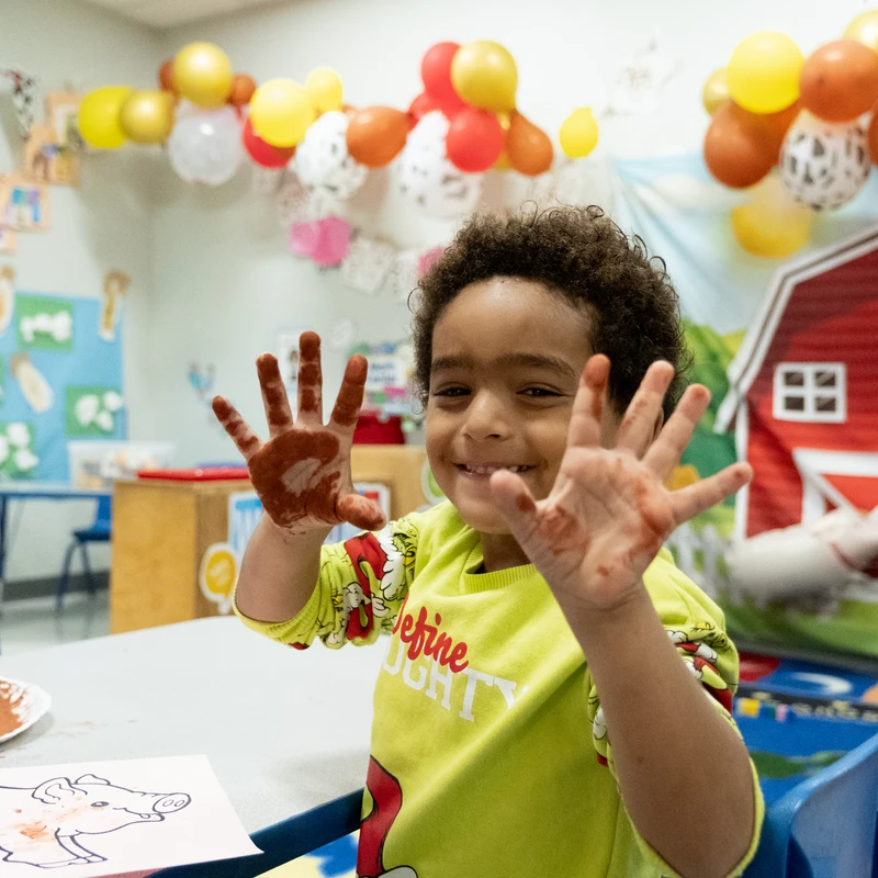 A smiling young child with curly hair proudly shows off their hands covered in brown paint while sitting at a table in a colorfully decorated classroom.