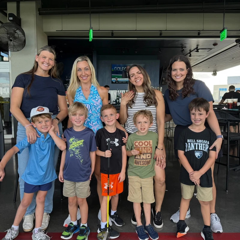 A group of four smiling women stand behind five young boys, posing for a photo at a covered driving range. One boy holds a golf club.
