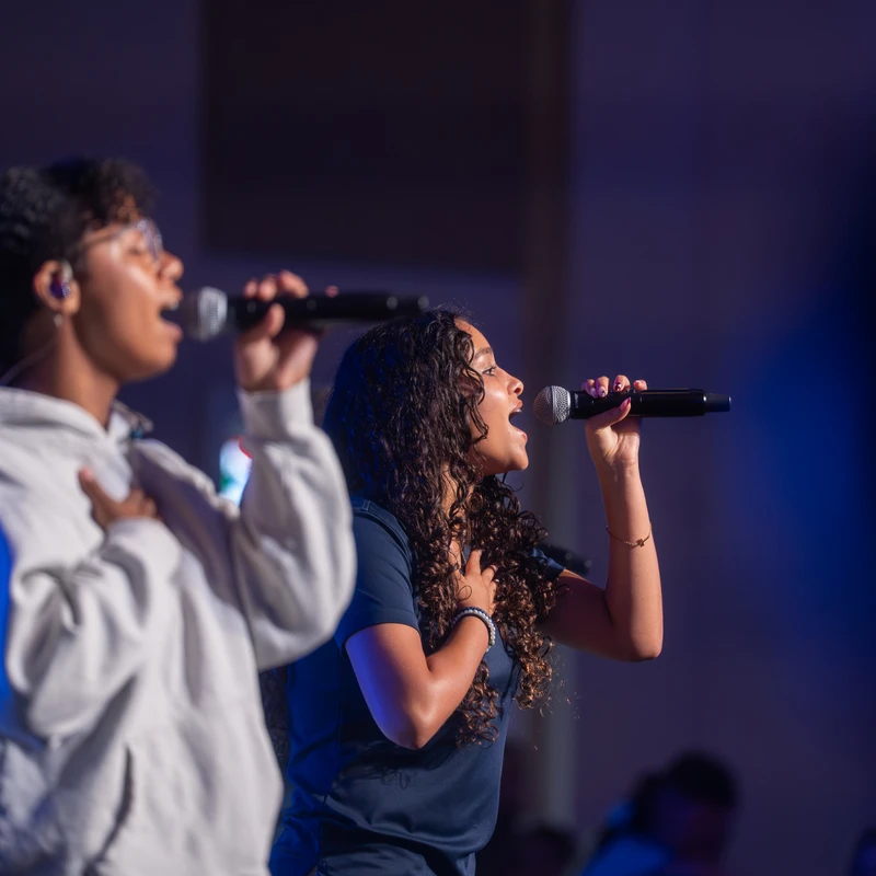 Two young women sing into microphones on a dimly lit stage. The woman in focus has curly hair and her hand on her chest. The other woman is slightly blurred.