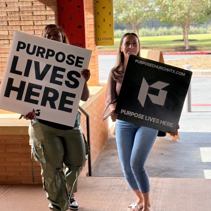 Two women stand outside a brick building, smiling and holding signs that read 'PURPOSE LIVES HERE' for Purpose Church.