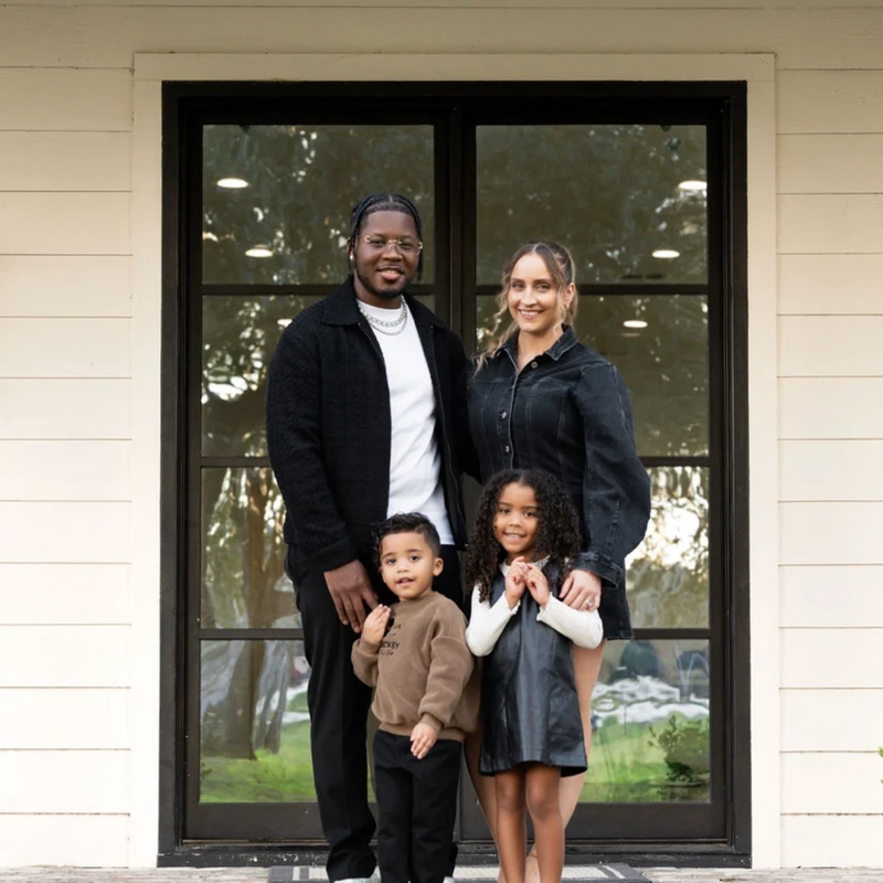 A smiling family of four, including a man, a woman, a little girl, and a little boy, stand together for a portrait on the brick porch of their house.