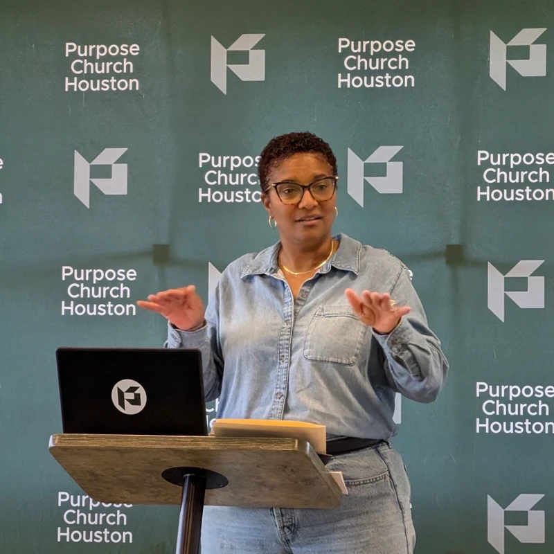 A woman with short hair and glasses wearing a denim outfit speaks from a podium. The background is a green banner for 'Purpose Church Houston'.