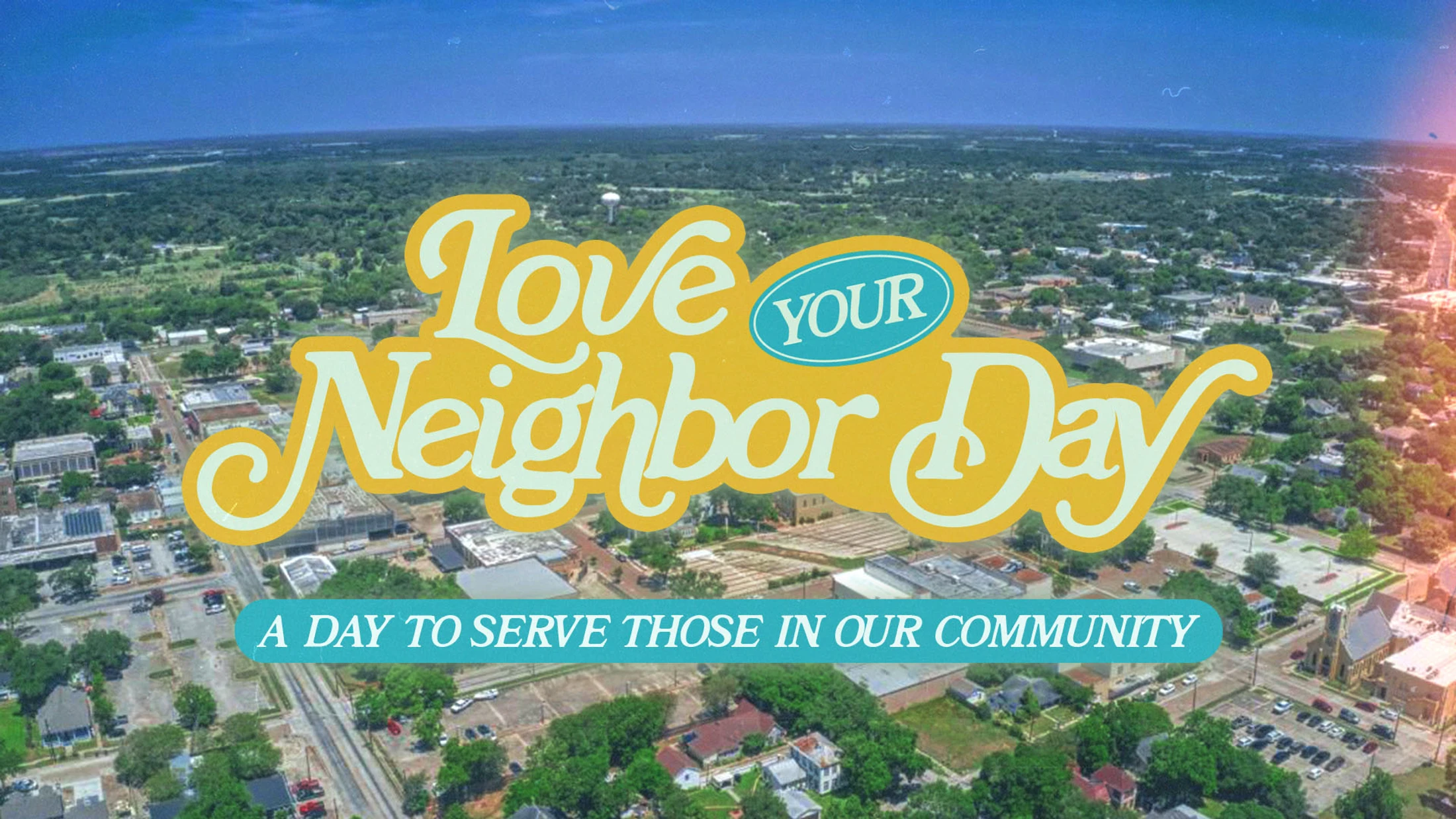 An aerial view of a small town with a graphic overlay that reads 'Love Your Neighbor Day, a day to serve those in our community.'