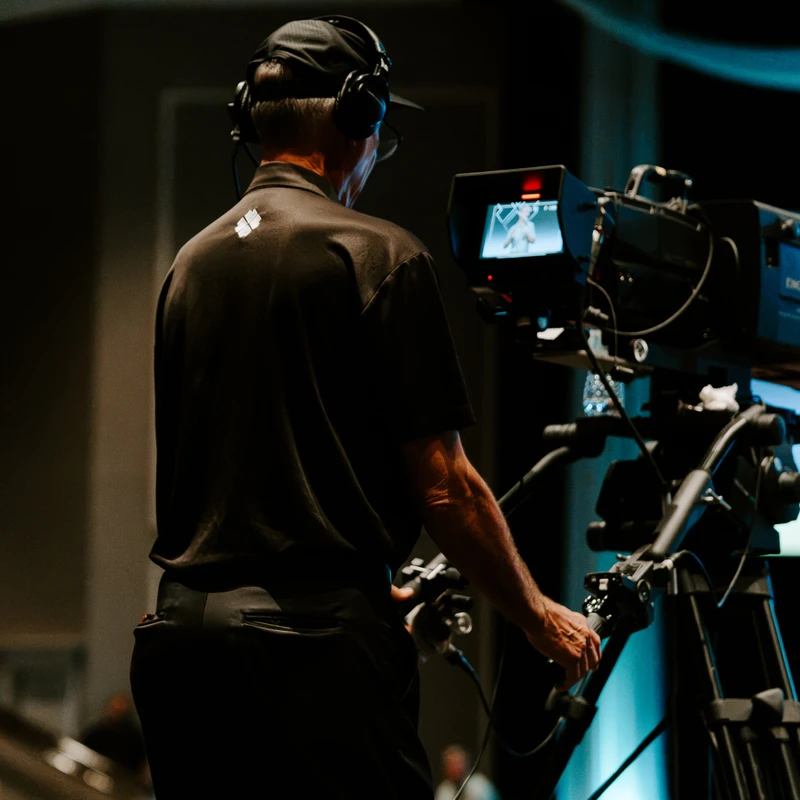 A camera operator, seen from behind, wears headphones and films a live event with a large professional video camera on a tripod in a dimly lit venue.