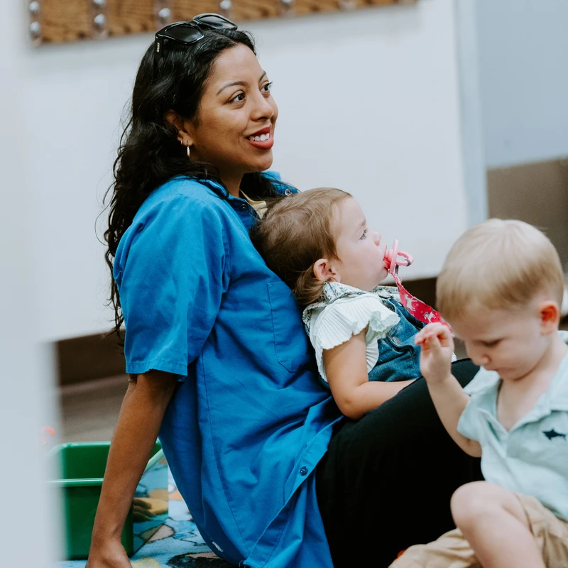 A woman in a blue shirt smiles while sitting on a colorful floor mat. A toddler girl with a pacifier rests on her lap, and a boy plays beside them.