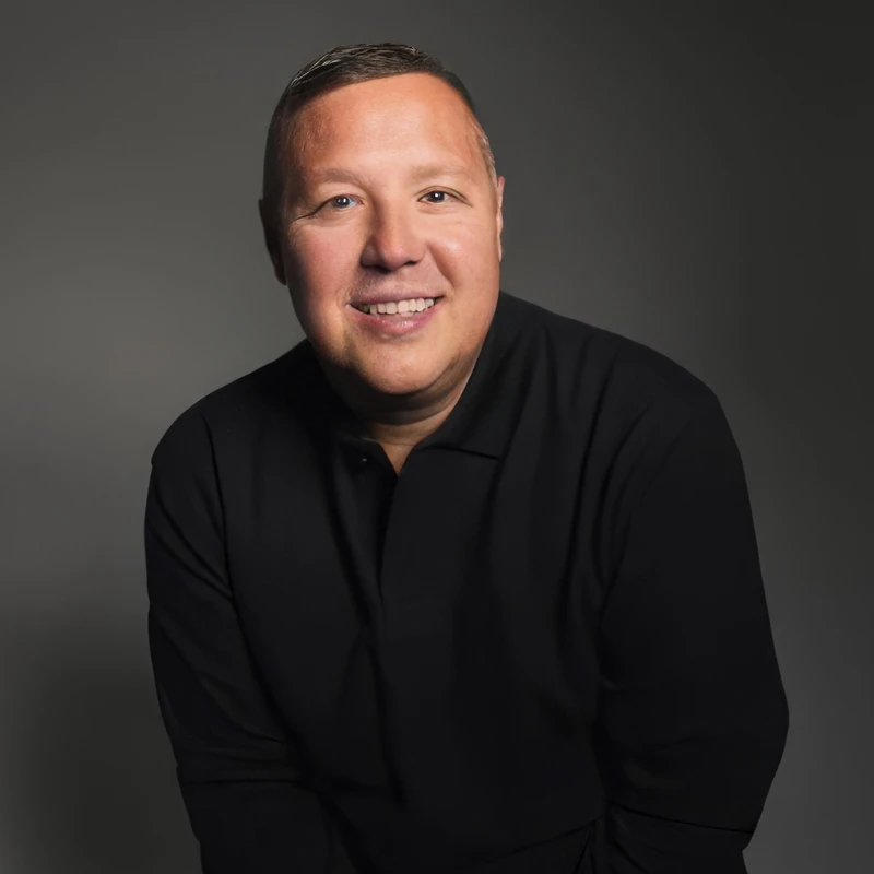 A studio portrait of a smiling middle-aged man with short brown hair, wearing a black polo shirt against a solid dark gray background.