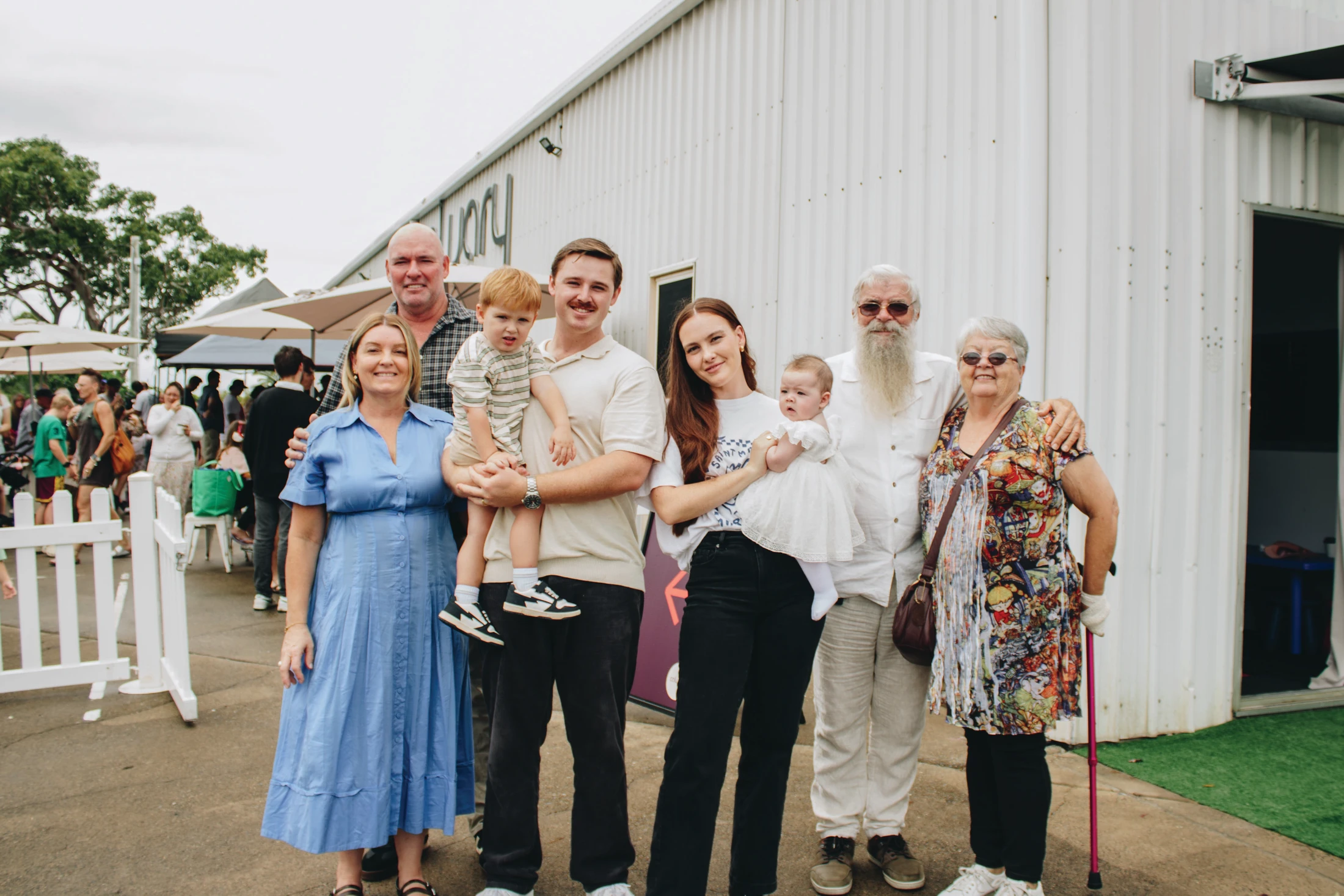 A large family with three generations, including grandparents, parents, a toddler, and a baby, posing for a group photo outdoors in front of a white building.
