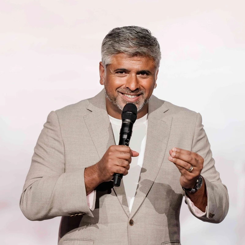 A man with graying hair and a beard smiles warmly while speaking into a microphone. He wears a light-colored suit jacket and gestures with his other hand.