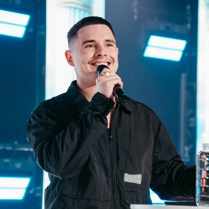 A young man with short dark hair and a mustache smiles while holding a microphone on a stage with bright blue and white lights in the background.