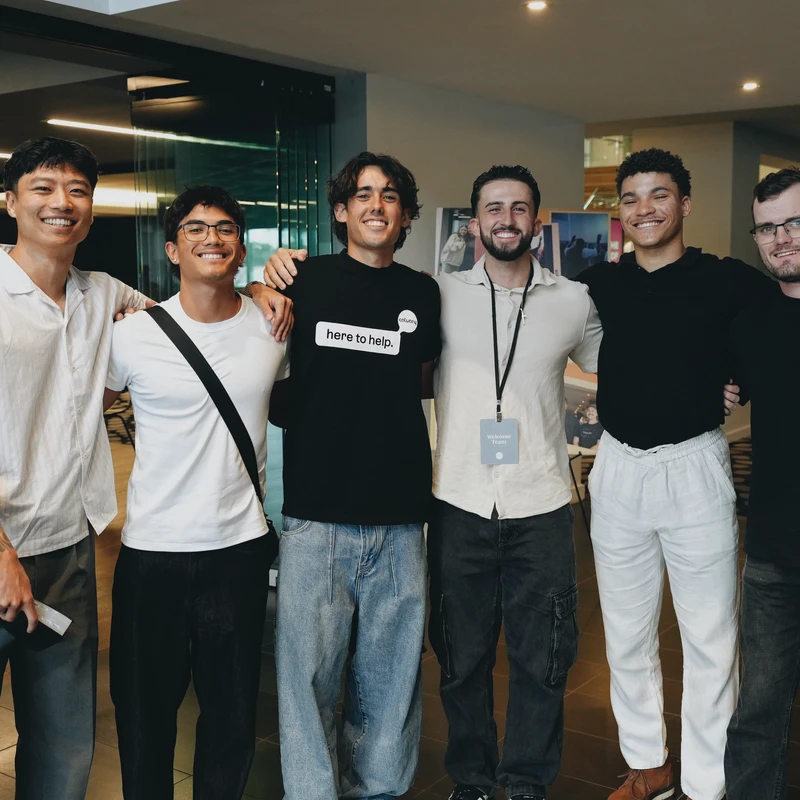 A group of six diverse young men stand side-by-side in a hallway with their arms around each other, smiling warmly for the camera.