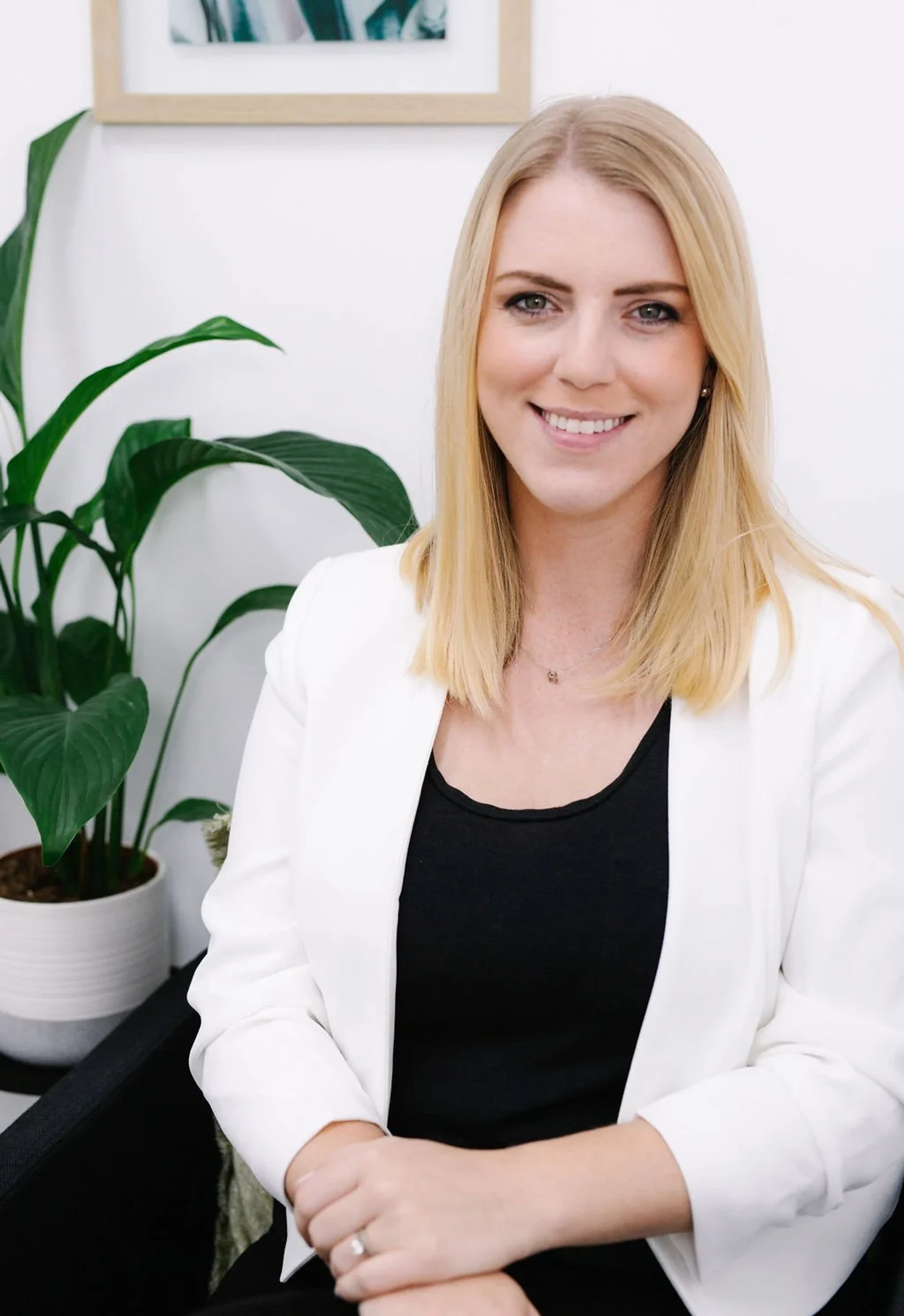 A professional headshot of a smiling blonde woman wearing a white blazer and black top. She is sitting in a chair with a green plant in the background.
