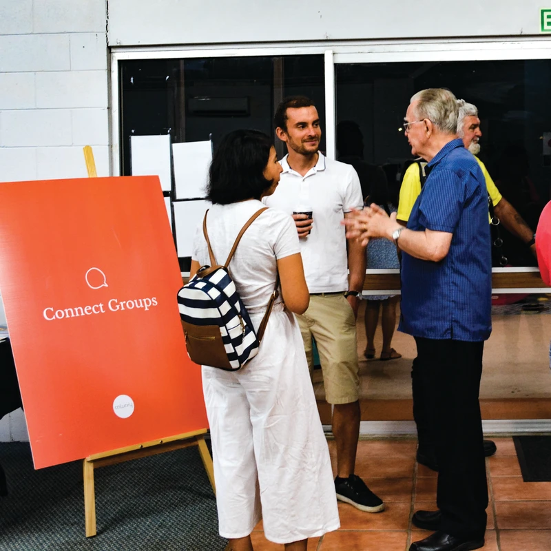 A diverse group of people stand and talk near an orange sign on an easel that reads 'Connect Groups' at an indoor gathering.