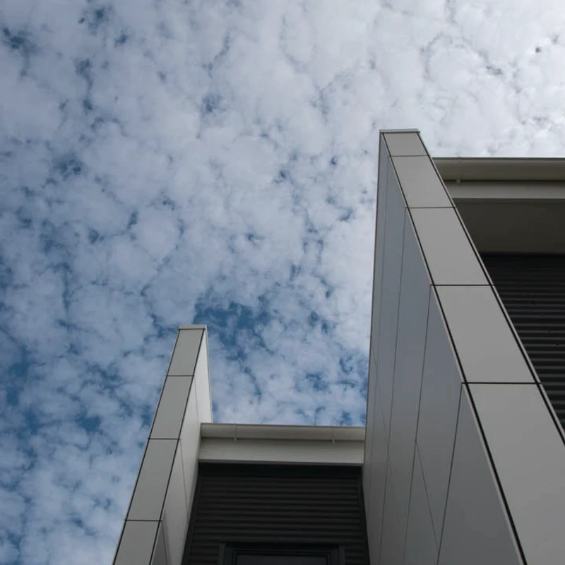 A low-angle shot of a modern building with angular gray panels and dark siding, set against a bright blue sky filled with scattered white clouds.