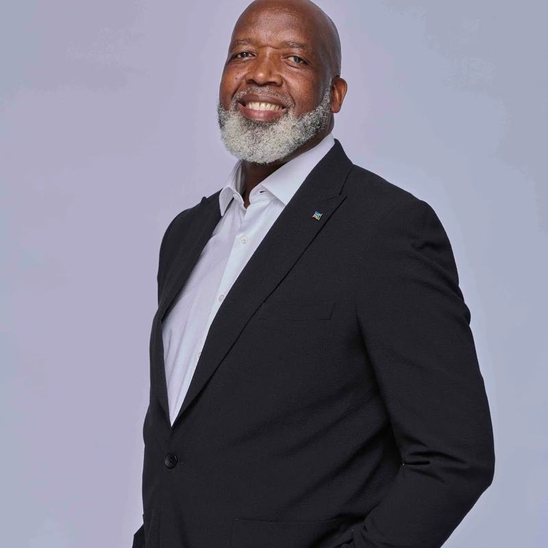 A smiling bald Black man with a full gray beard poses in a black suit and white shirt against a plain light gray background, with his hands in his pockets.