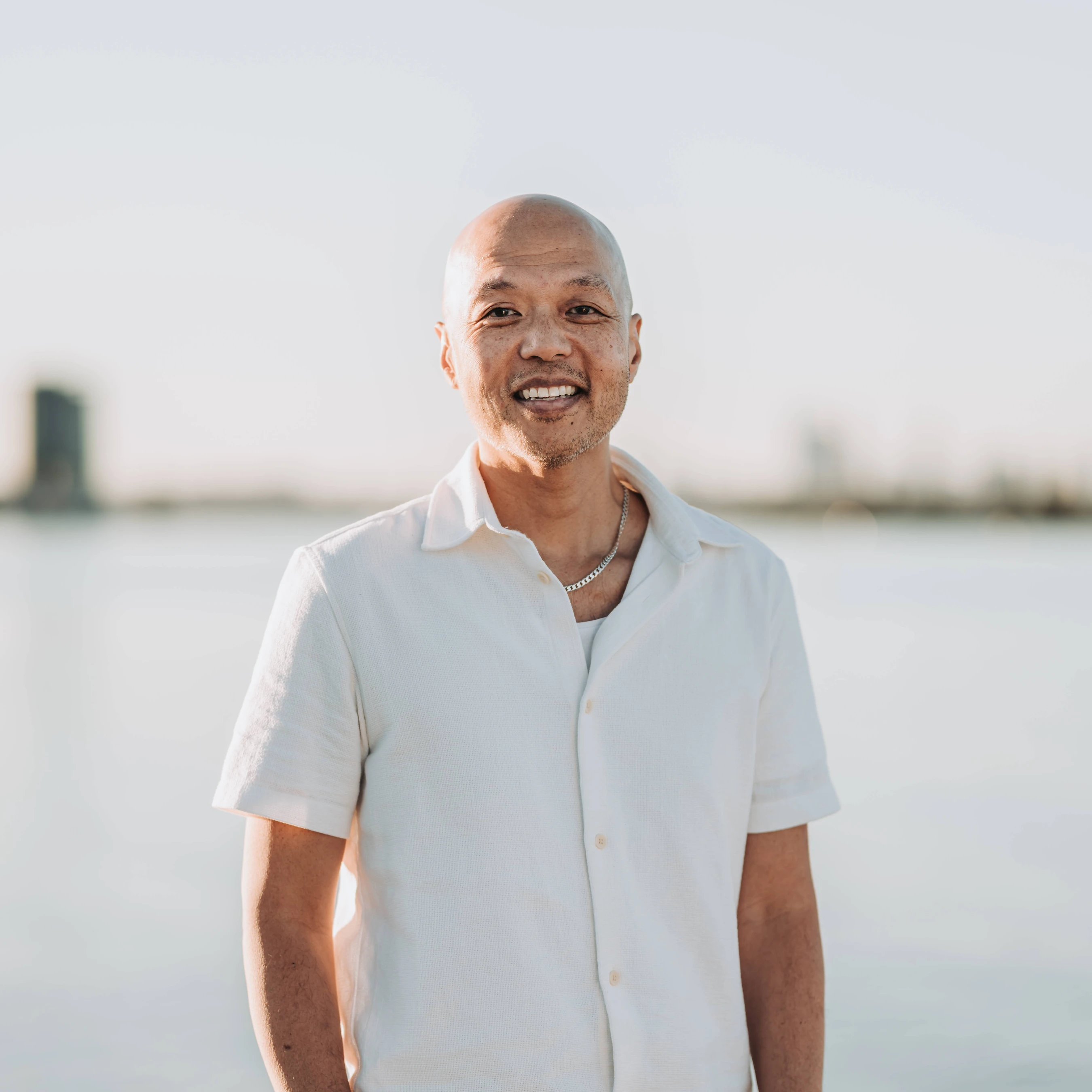 A middle-aged Asian man with a shaved head smiles at the camera. He wears a white shirt and stands outdoors with water and a city skyline in the background.