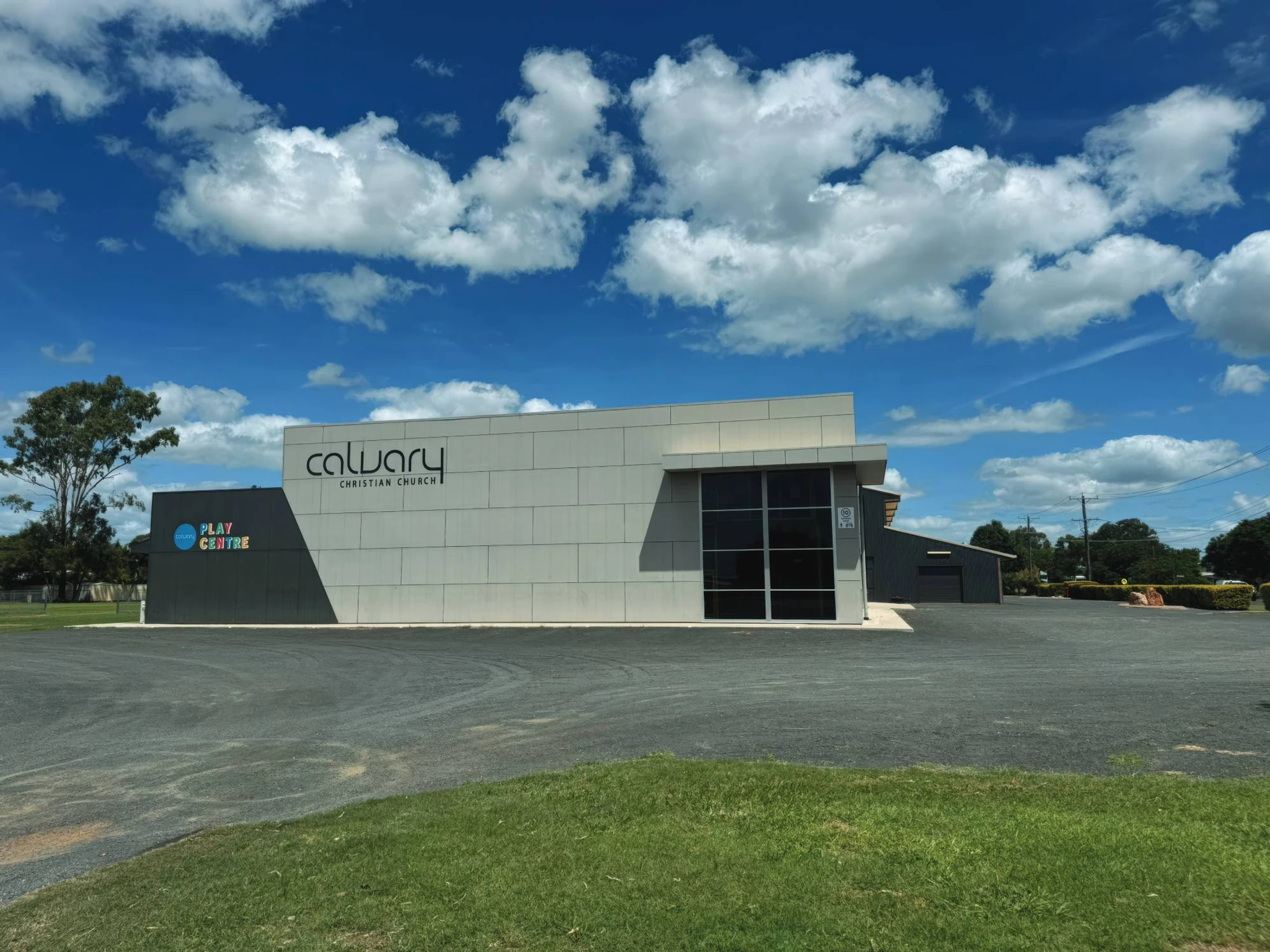 Exterior of the modern Calvary Christian Church and Play Centre. The building sits under a bright blue sky with fluffy white clouds, with a gravel lot in front.