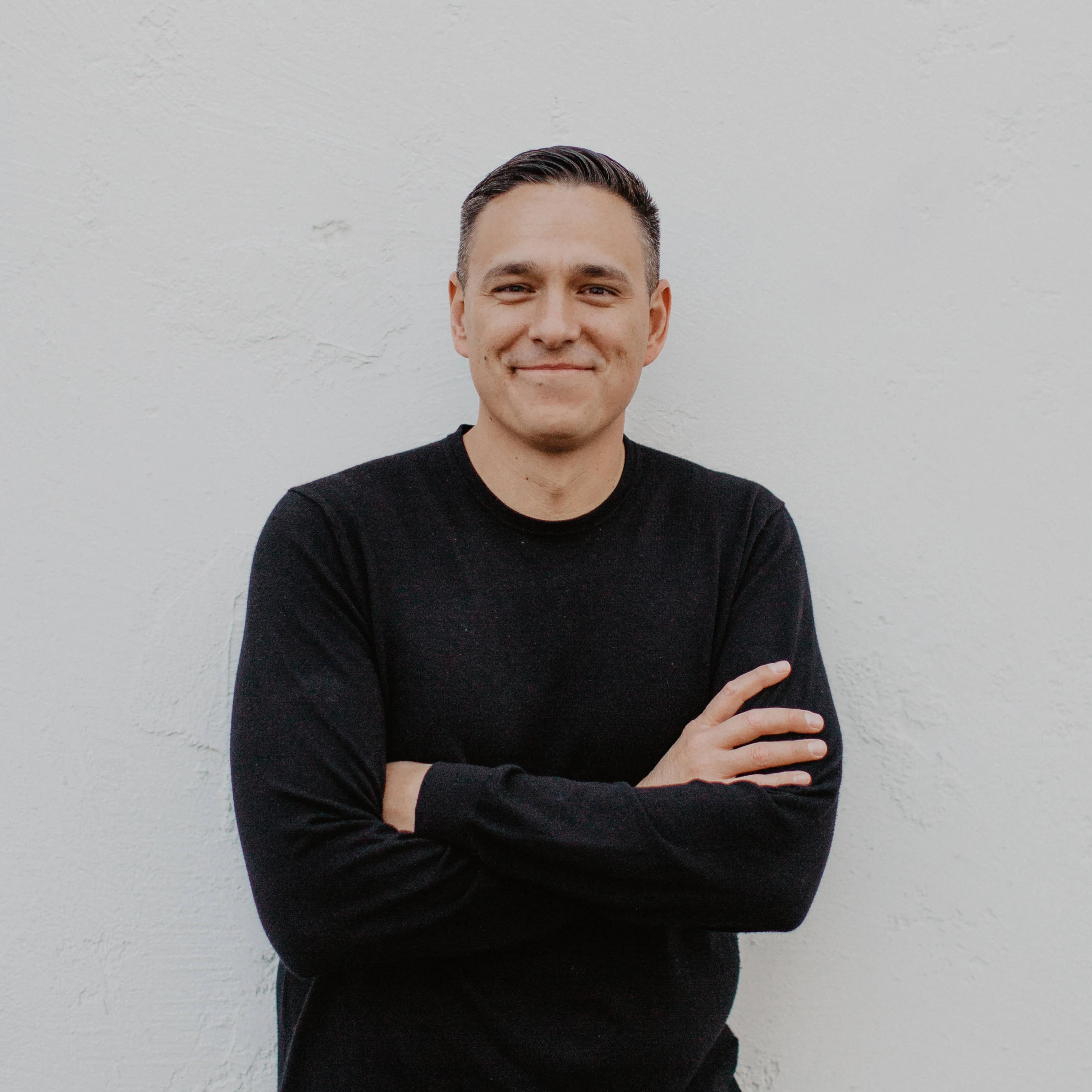 A smiling man with short dark hair, wearing a black sweater, stands in front of a plain white wall with his arms crossed.