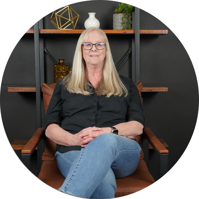 A woman with long blonde hair and glasses, wearing a black shirt and jeans, sits smiling in a brown leather armchair against a dark background.