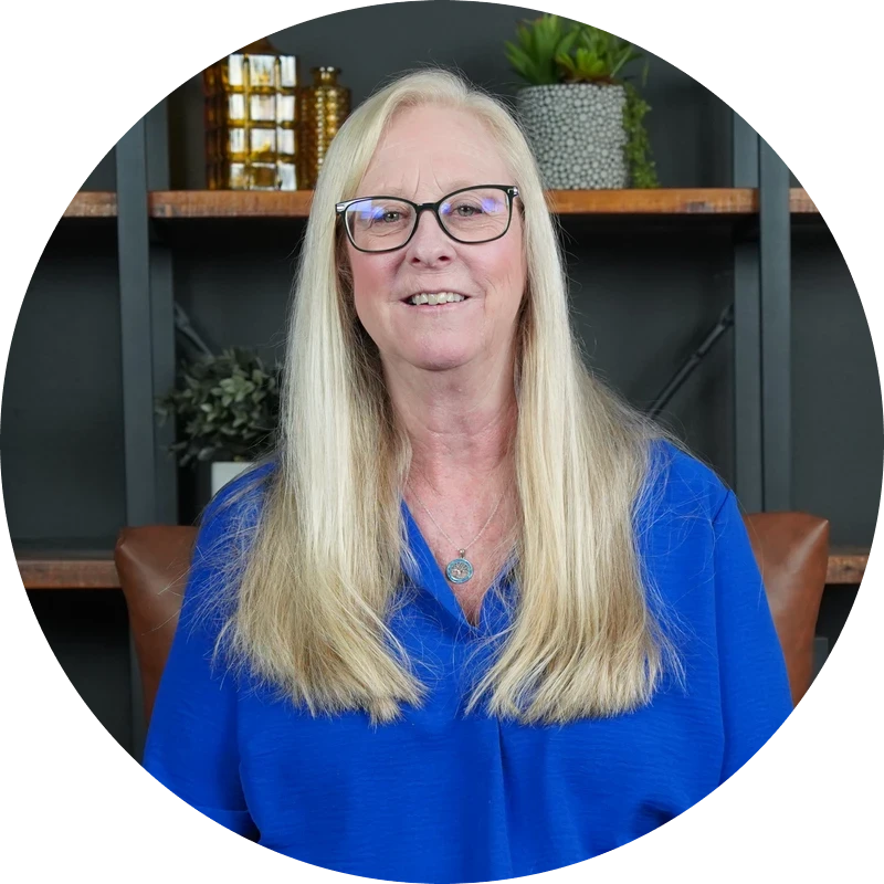 A circular portrait of a smiling woman with long blonde hair. She is wearing black glasses, a blue top, and a necklace, sitting in front of a bookshelf.