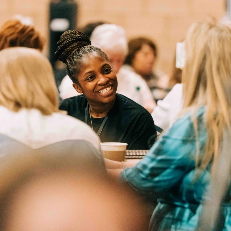 A Black woman with her hair in a braided bun smiles warmly while talking with other people at a table during a conference or workshop.