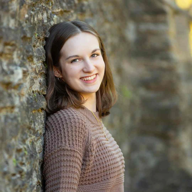 A young woman with shoulder-length brown hair and a brown knit sweater smiles warmly at the camera while leaning against a rustic stone wall outdoors.