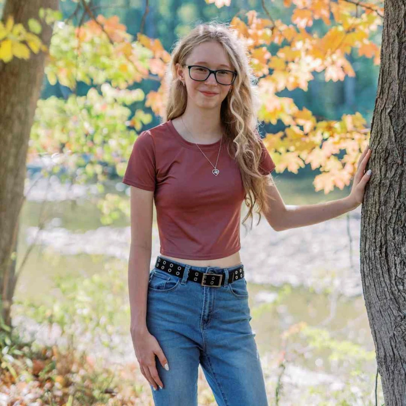 A young woman with long blonde hair and glasses smiles, leaning against a tree. She wears a rust-colored crop top and jeans in a wooded area in autumn.