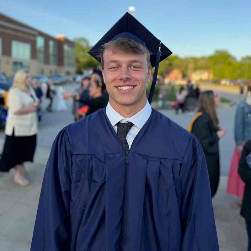 A smiling young man poses for a photo outdoors, wearing a dark blue graduation cap and gown over a white shirt and tie.
