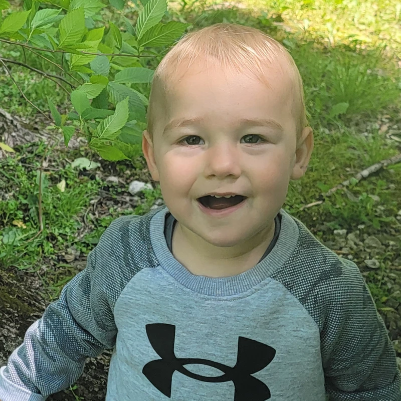 A close-up portrait of a young boy with blond hair, smiling happily at the camera while sitting outdoors in front of green leaves and grass.