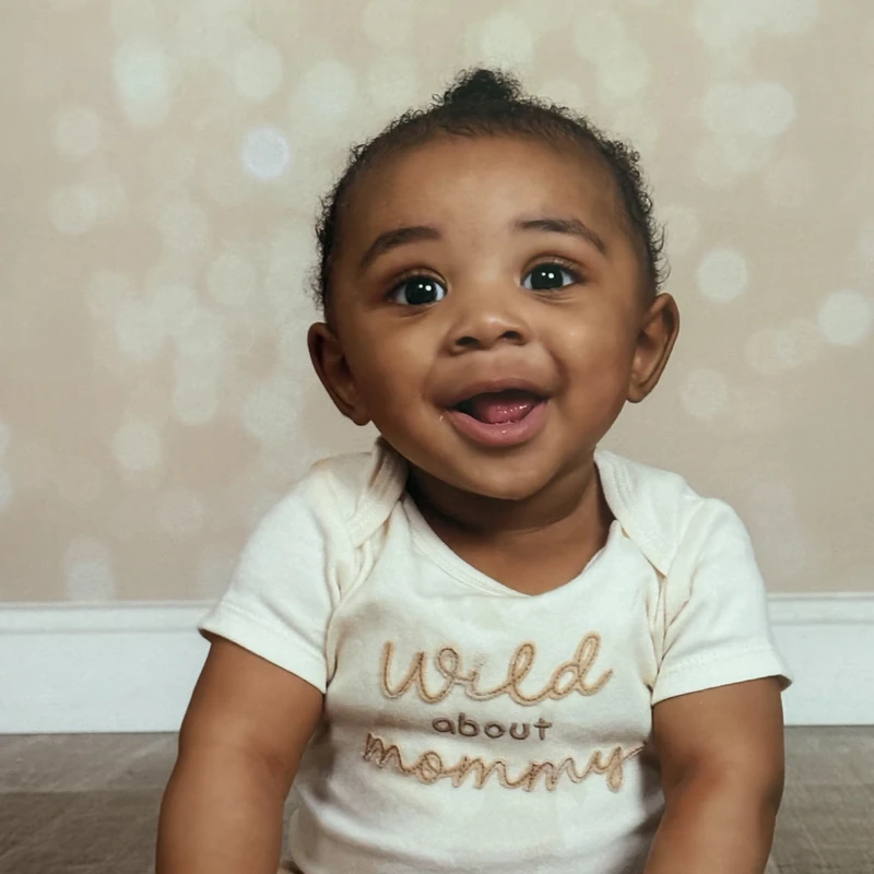 A close-up portrait of a smiling Black baby with big brown eyes, wearing a cream onesie that says 'Wild about mommy' against a light, blurry background.