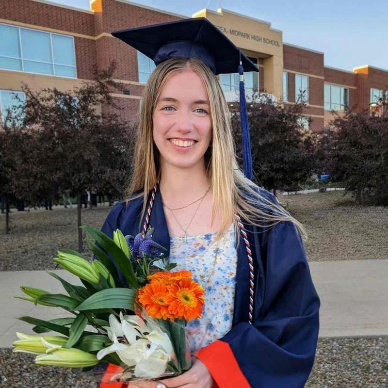 A smiling young woman in a graduation cap and gown holds a colorful bouquet of flowers while standing in front of Midpark High School.