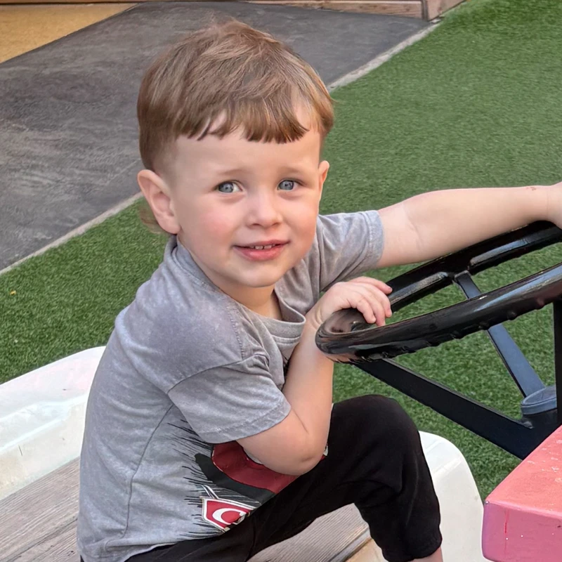 A young boy with blue eyes and a brown mullet haircut sits in a toy vehicle on a playground. He is smiling and holding the steering wheel.