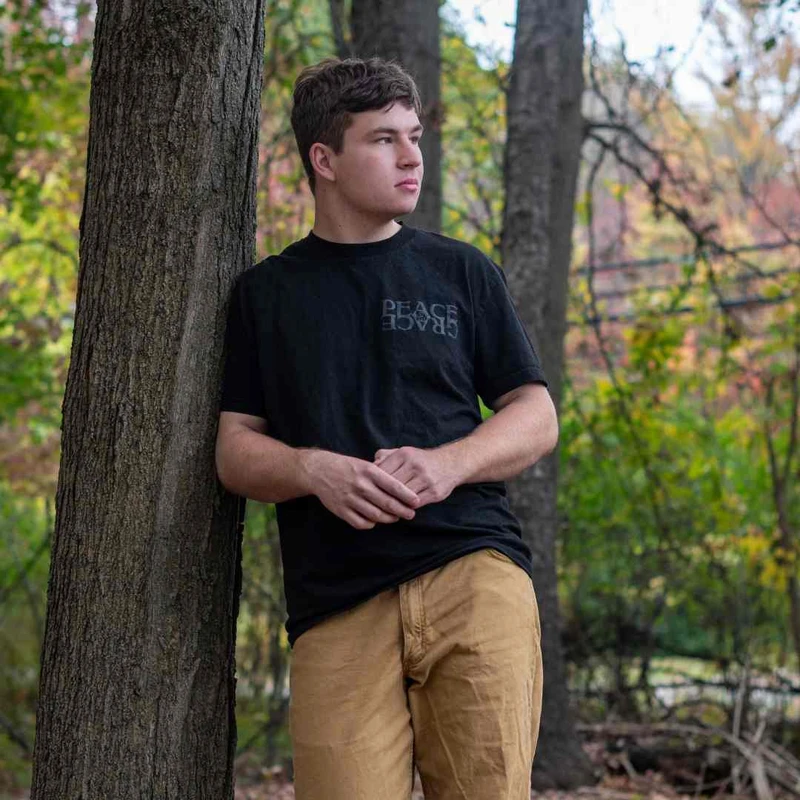A young man with short brown hair leans against a tree in a forest. He is wearing a black t-shirt with text and tan pants, looking off to the side.